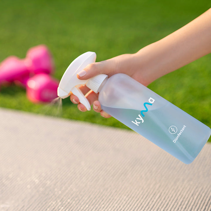 Hand spraying Kyma Disinfectant cleaner on a yoga mat outdoors, with pink dumbbells in the background.