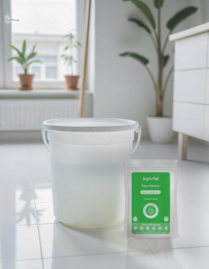 Kyma Floor Cleaner Forest Dew variant placed beside a bucket of cleaning water on a spotless white floor in a bright, plant-filled room showcasing an eco-friendly, powder-based cleaner for naturally fresh and sparkling floors.