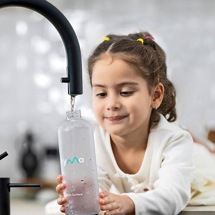 Person filling a Kyma Multi-Surface Cleaner spray bottle with water from a modern black kitchen faucet, preparing the eco-friendly cleaning solution.