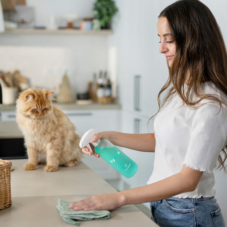 Woman cleaning a kitchen counter with a Kyma Multi-Surface cleaner spray while a fluffy orange cat sits nearby.