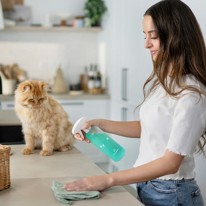 Woman cleaning a kitchen counter with a Kyma Multi-Surface cleaner spray while a fluffy orange cat sits nearby.