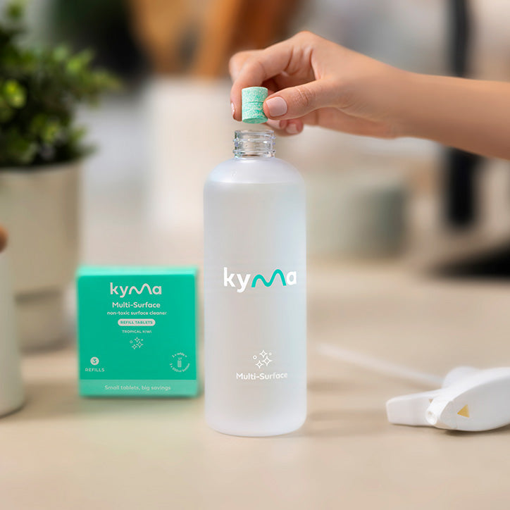 Person dropping a Kyma cleaning tablet into a reusable bottle of water, with the Kyma Multi-Surface cleaner box and spray nozzle on the counter.