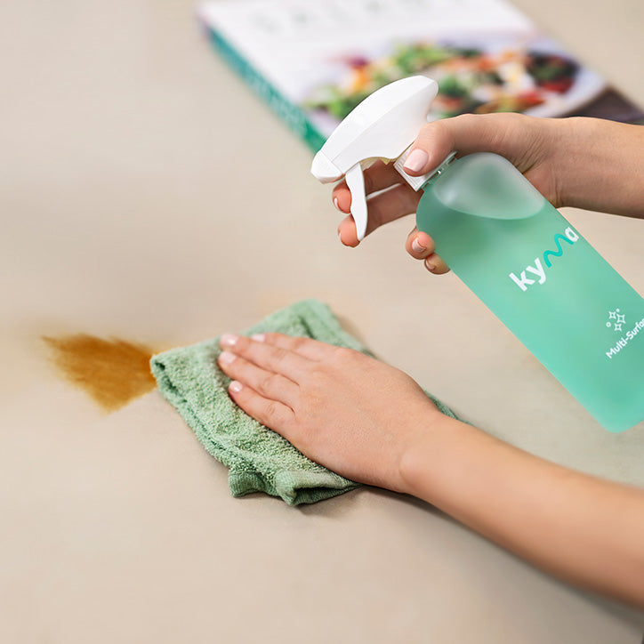 Person cleaning a stained countertop using a Kyma Multi-Surface Cleaner spray bottle and a green microfiber cloth, demonstrating the product’s powerful stain removal capability.