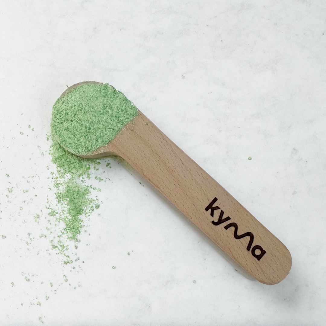 Kyma 10g wooden scoop filled with green cleaning powder, resting on a white surface with some powder scattered around.