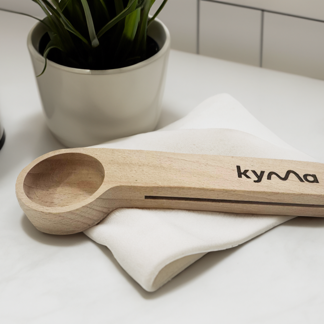 Kyma 10g wooden measuring scoop placed on a soft white cloth beside a potted plant crafted for precise and sustainable home use.