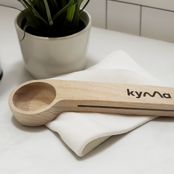 Kyma 10g wooden measuring scoop placed on a soft white cloth beside a potted plant crafted for precise and sustainable home use.