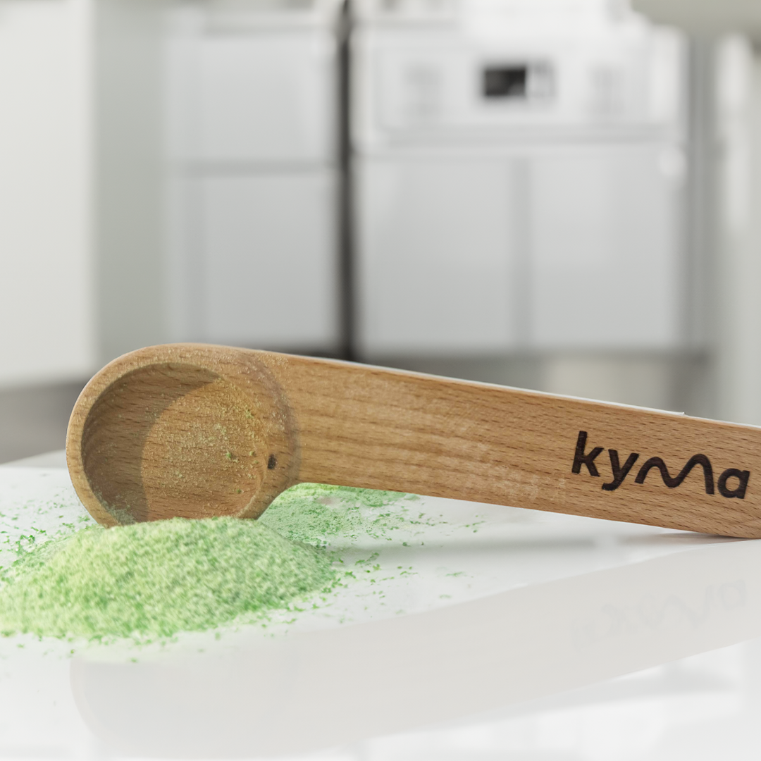Kyma 6 gram sustainable scoop resting on a green powdered substance, with a kitchen setting in the background.
