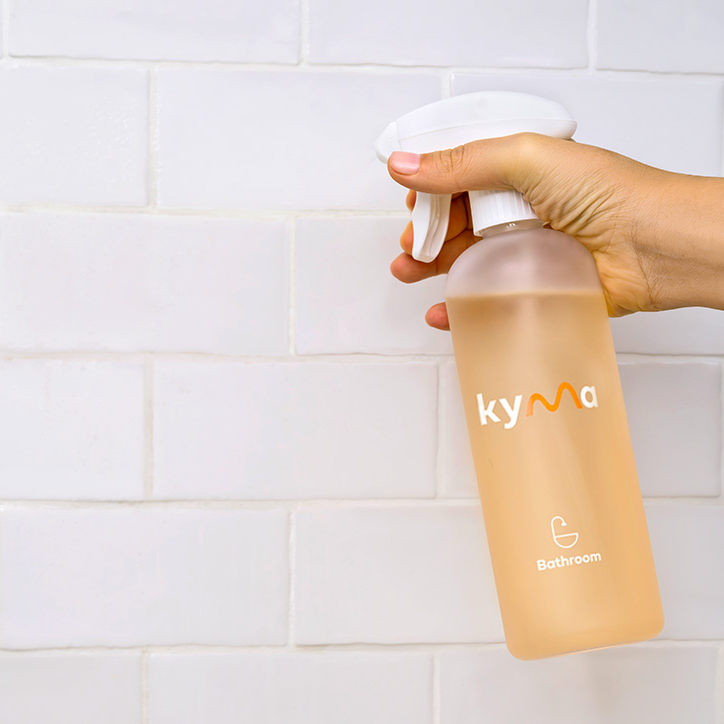 Hand holding a Kyma Bathroom Cleaner spray bottle against a tiled wall.