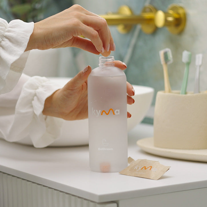 Person dropping a Kyma bathroom cleaning tablet into a refillable spray bottle filled with water on a bathroom counter.