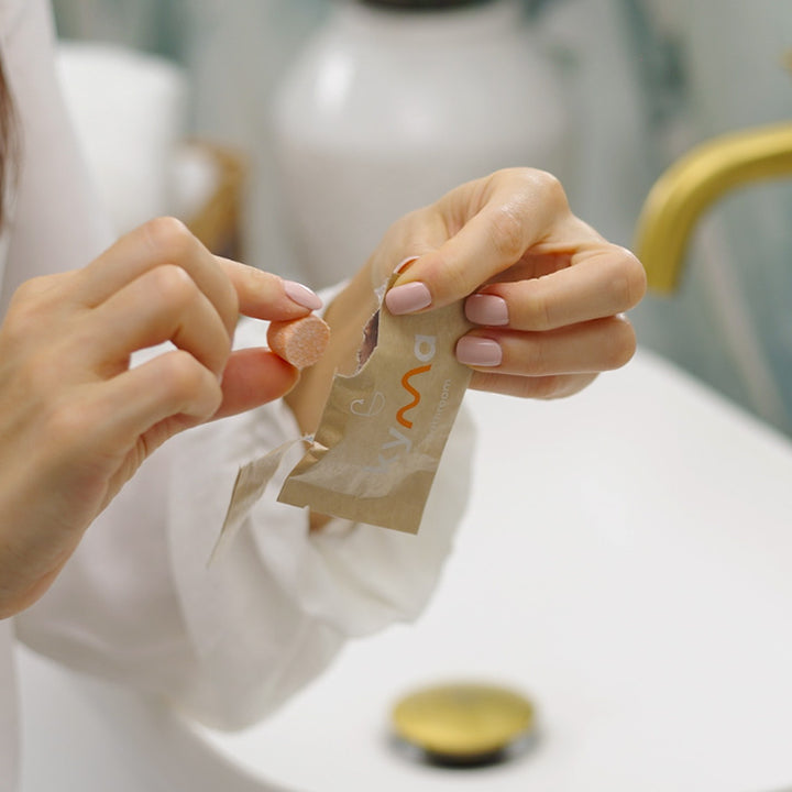 Person unwrapping a Kyma bathroom cleaning tablet from its paper packaging near a sink.