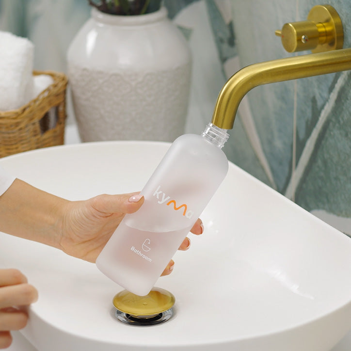 Hand filling a Kyma Bathroom Cleaner bottle with water from a gold faucet.