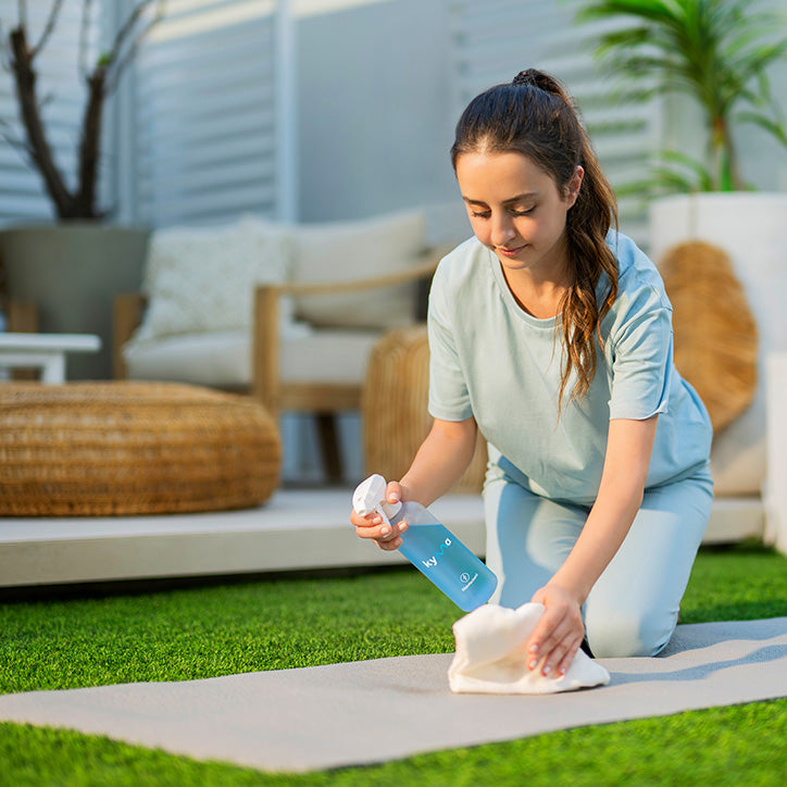 A woman sprays a Kyma disinfectant bottle while cleaning a mat outdoors on the grass.