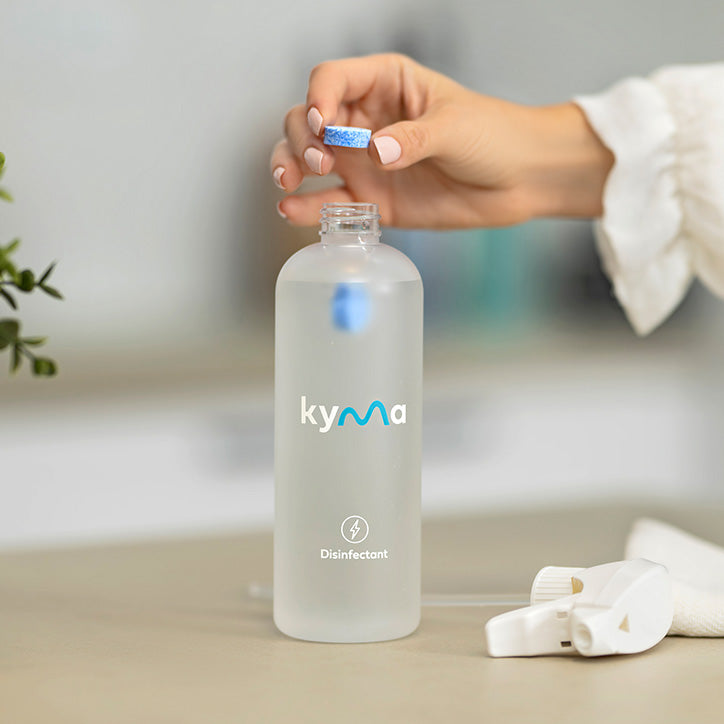 A person drops a blue Kyma disinfectant tablet into a clear refillable spray bottle labeled “Disinfectant” on a countertop.
