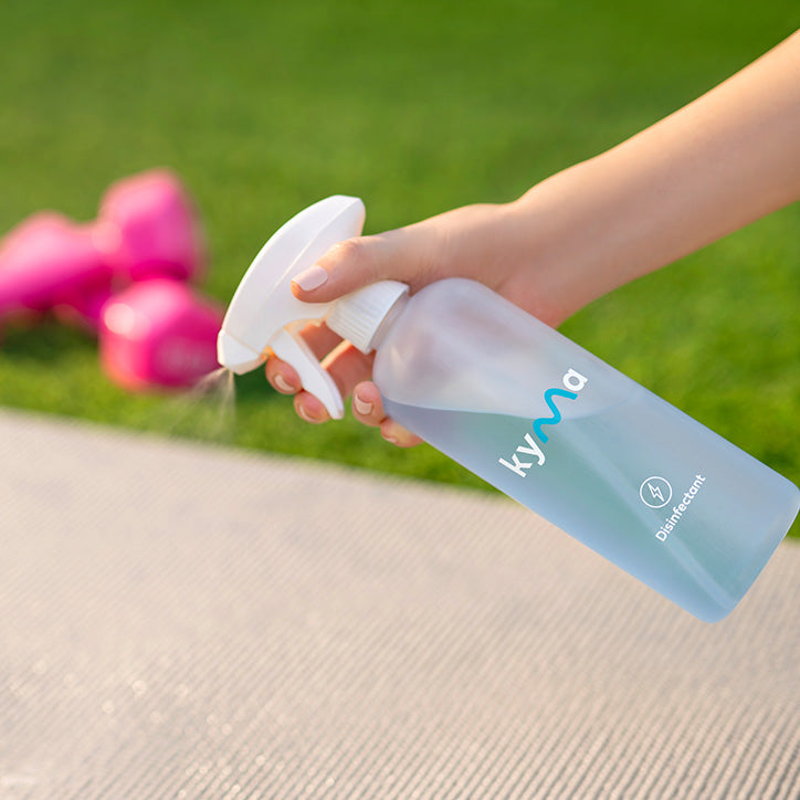 Hand spraying Kyma Disinfectant cleaner on a yoga mat outdoors, with pink dumbbells in the background.