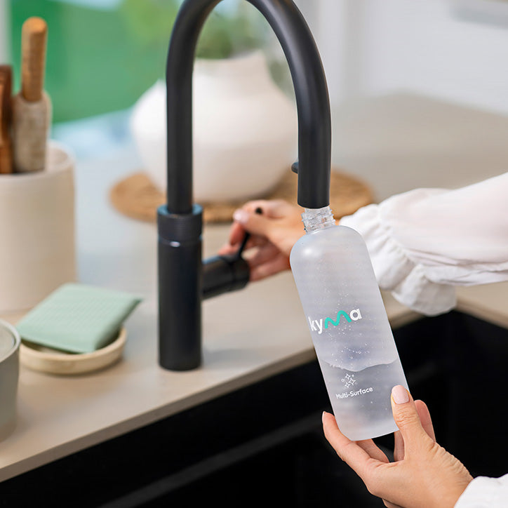 Person refilling a Kyma multi-surface cleaner bottle with water at a kitchen sink.
