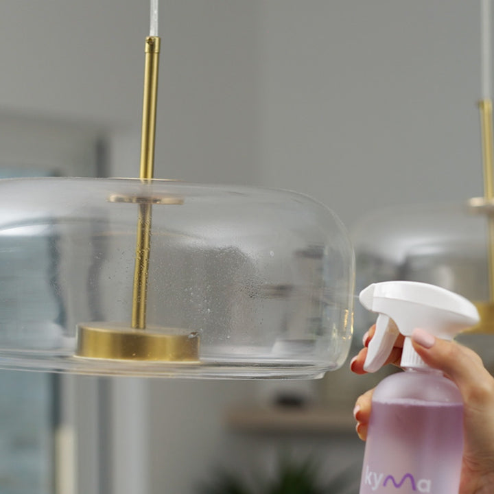 Person using Kyma glass cleaner spray to clean a transparent glass lampshade with a gold fixture, leaving a clear and streak-free shine.