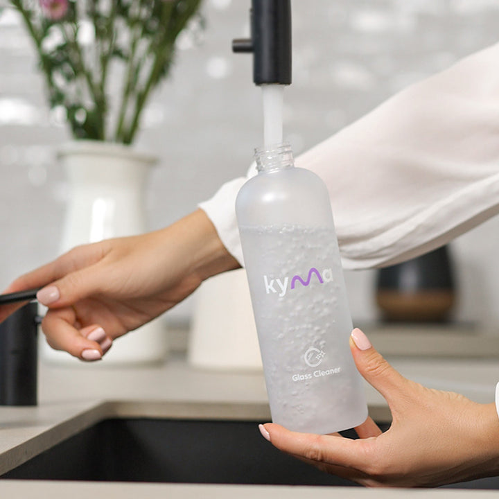 Person filling a Kyma Glass Cleaner reusable spray bottle with water from a kitchen tap, preparing for eco-friendly cleaning.