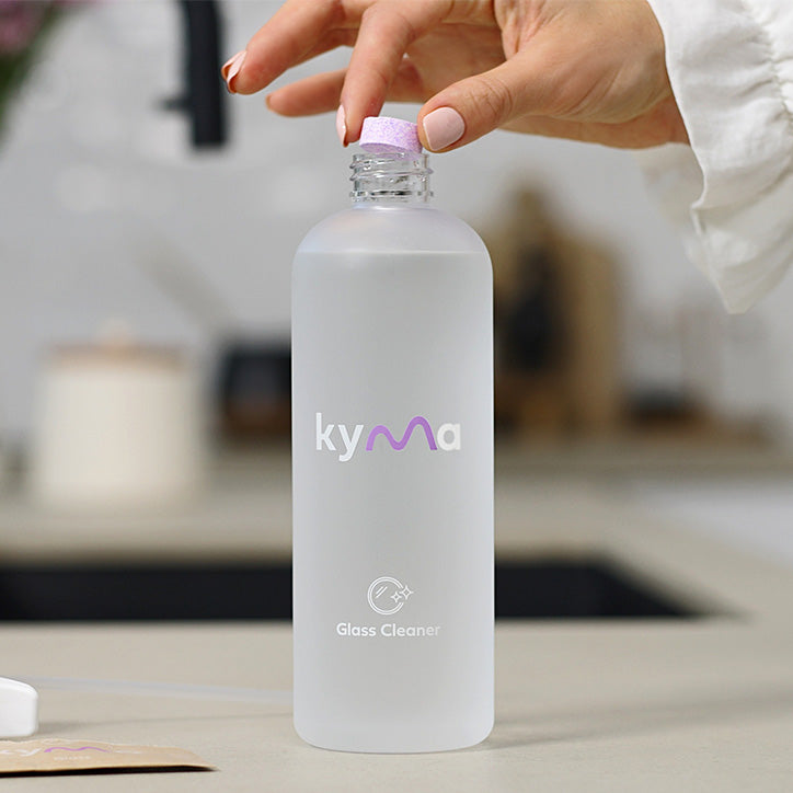Hand dropping a Kyma glass cleaner refill tablet into a reusable spray bottle filled with water, demonstrating the eco-friendly cleaning refill process.