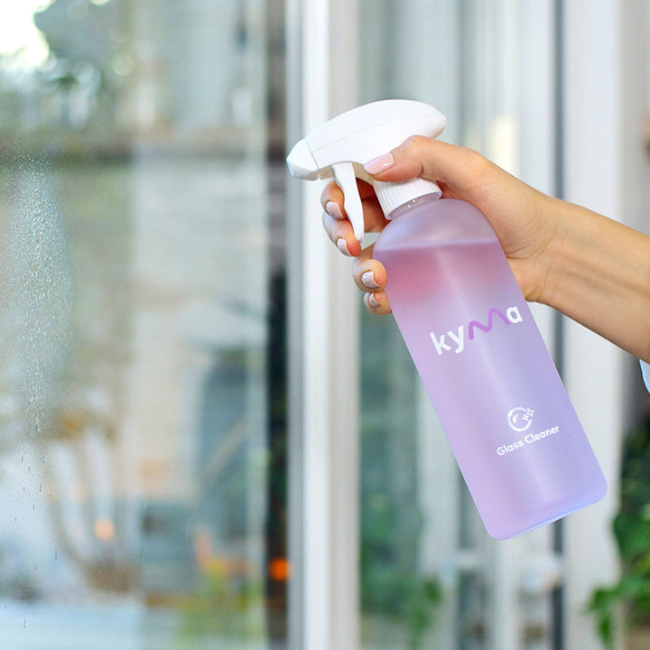 Person spraying Kyma Glass Cleaner on a window for a streak-free shine, showing a lavender-colored reusable bottle and clear surface reflection.