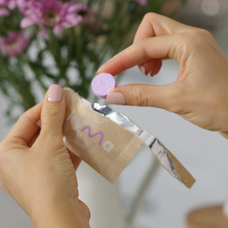 Person holding a Kyma glass cleaner refill tablet and unwrapping it from eco-friendly packaging, ready to dissolve in water for use.