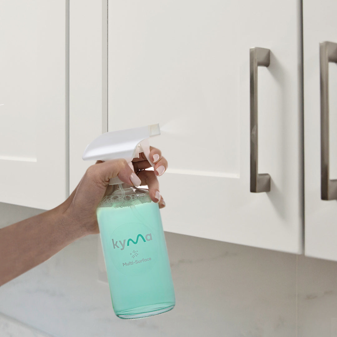 A person using the Kyma Multi-Surface Cleaner spray bottle with light blue liquid to clean white kitchen cabinets, showcasing an eco-friendly and reusable cleaning solution.