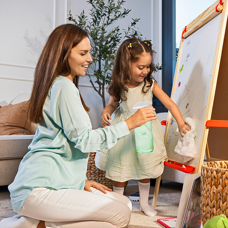 A mother and daughter cleaning together using a Kyma Multi-Surface Cleaner spray bottle, wiping a whiteboard in a cozy living room highlighting safe, eco-friendly cleaning.