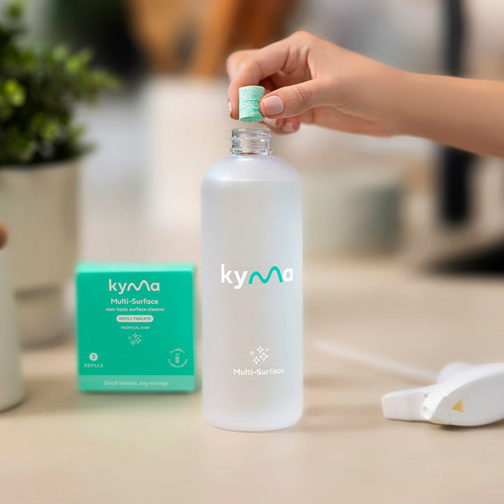 Person dropping a Kyma cleaning tablet into a reusable bottle of water, with the Kyma Multi-Surface cleaner box and spray nozzle on the counter.